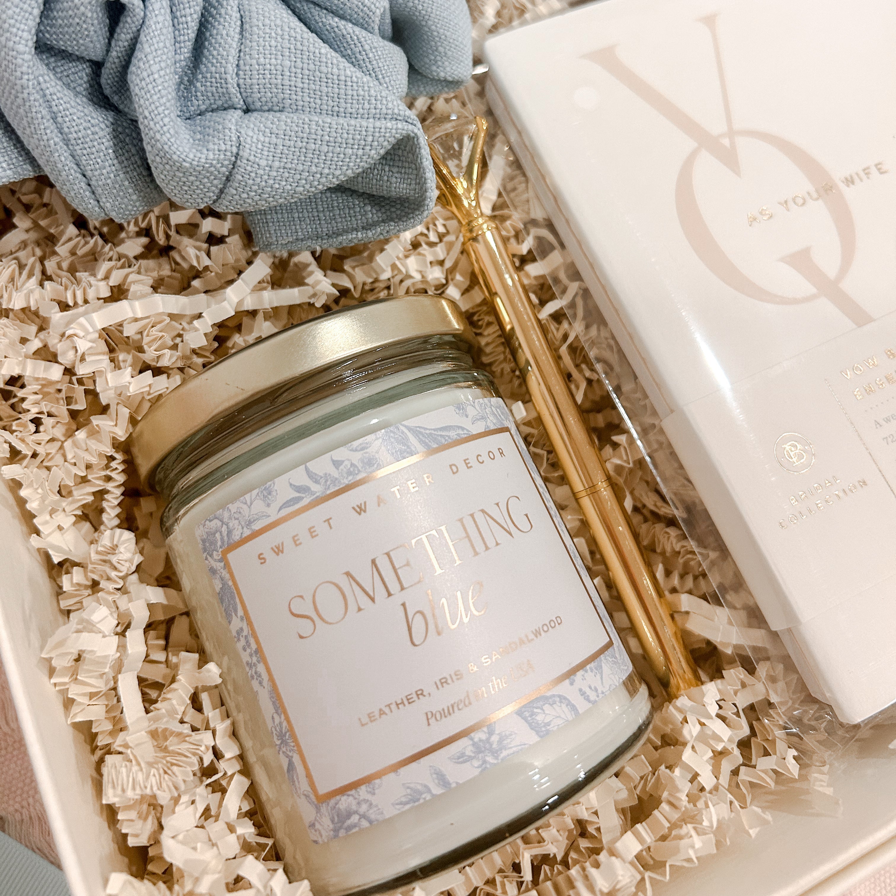 Close-up of the Something Blue Bride-to-Be Gift Box featuring a Something Blue soy candle, powder blue jumbo scrunchie, embroidered bride socks, gold diamond pen, and keepsake vow books.