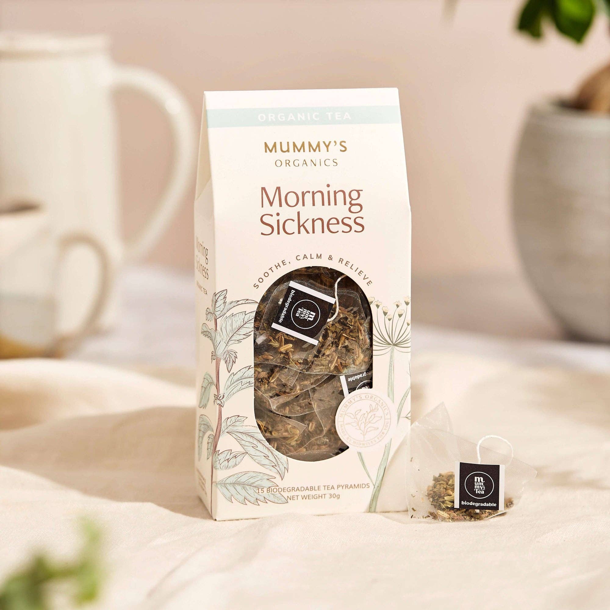 A package of Mummy's Organics Morning Sickness organic tea stands on a soft cream fabric surface, with a small tea bag placed beside it and a blurred mug and plant in the background.