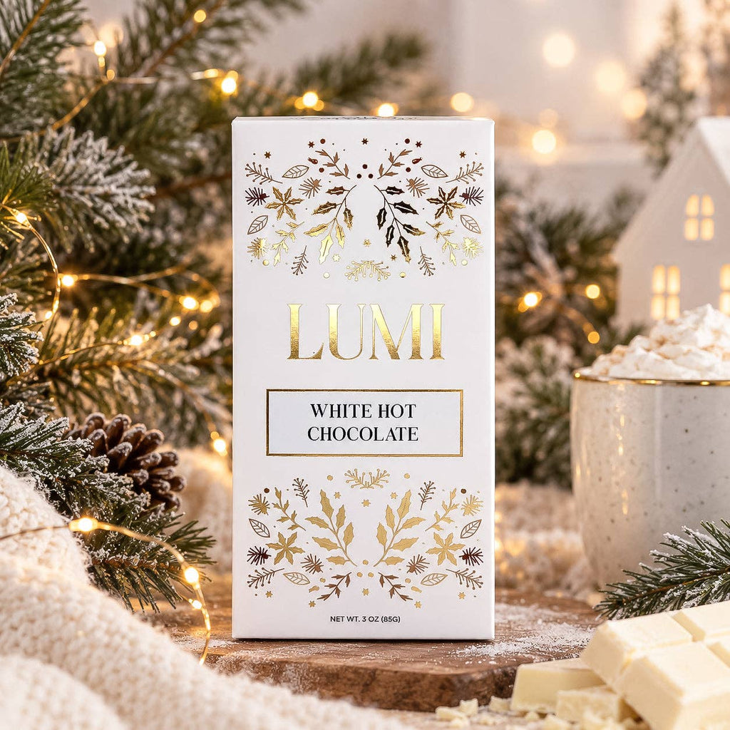 A boxed bar of LUMI White Hot Chocolate chocolate stands on a rustic wooden surface surrounded by holiday greenery, warm string lights, whipped cream, and chocolate pieces. The box is white with gold floral illustrations and the brand name LUMI in large gold letters.