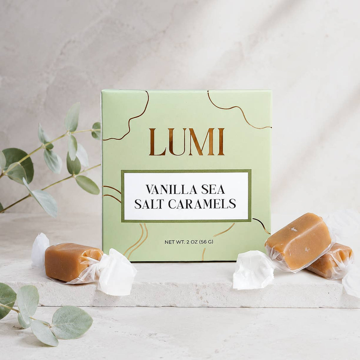 A box of LUMI Vanilla Sea Salt Caramels sits on a light stone surface with several wrapped caramel pieces scattered around it and eucalyptus leaves in the foreground.