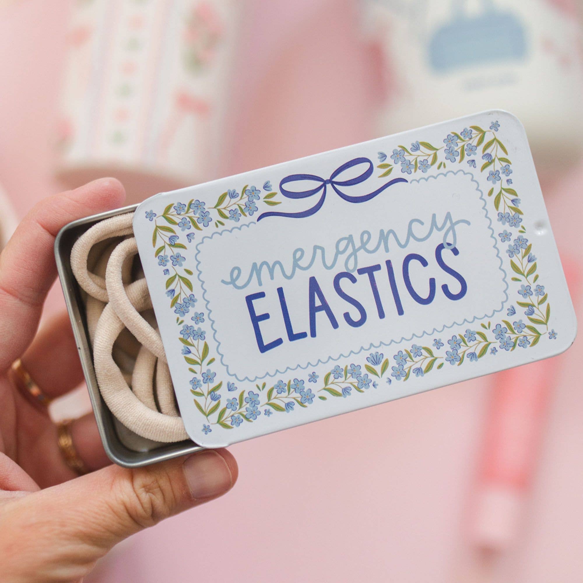 Small tin labeled 'emergency elastics' held by a hand against a blurred background