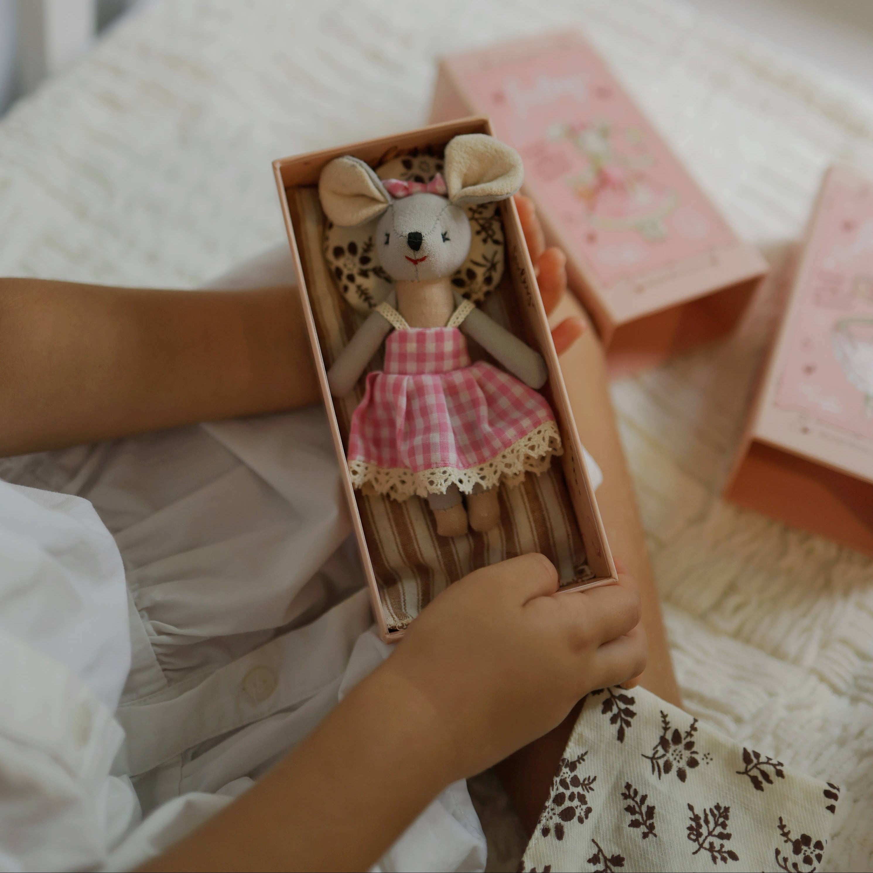 Child holding a small mouse doll in a box on a bed with pink books.