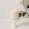 Candle in a jar labeled 'thank you!' on a small white stand with a blurred background