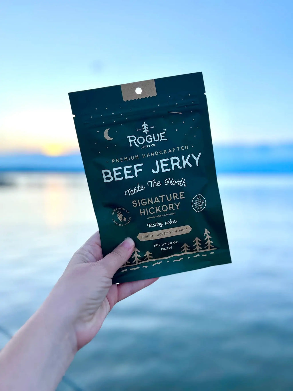 Hand holding a package of Rogue Beef Jerky against a scenic background