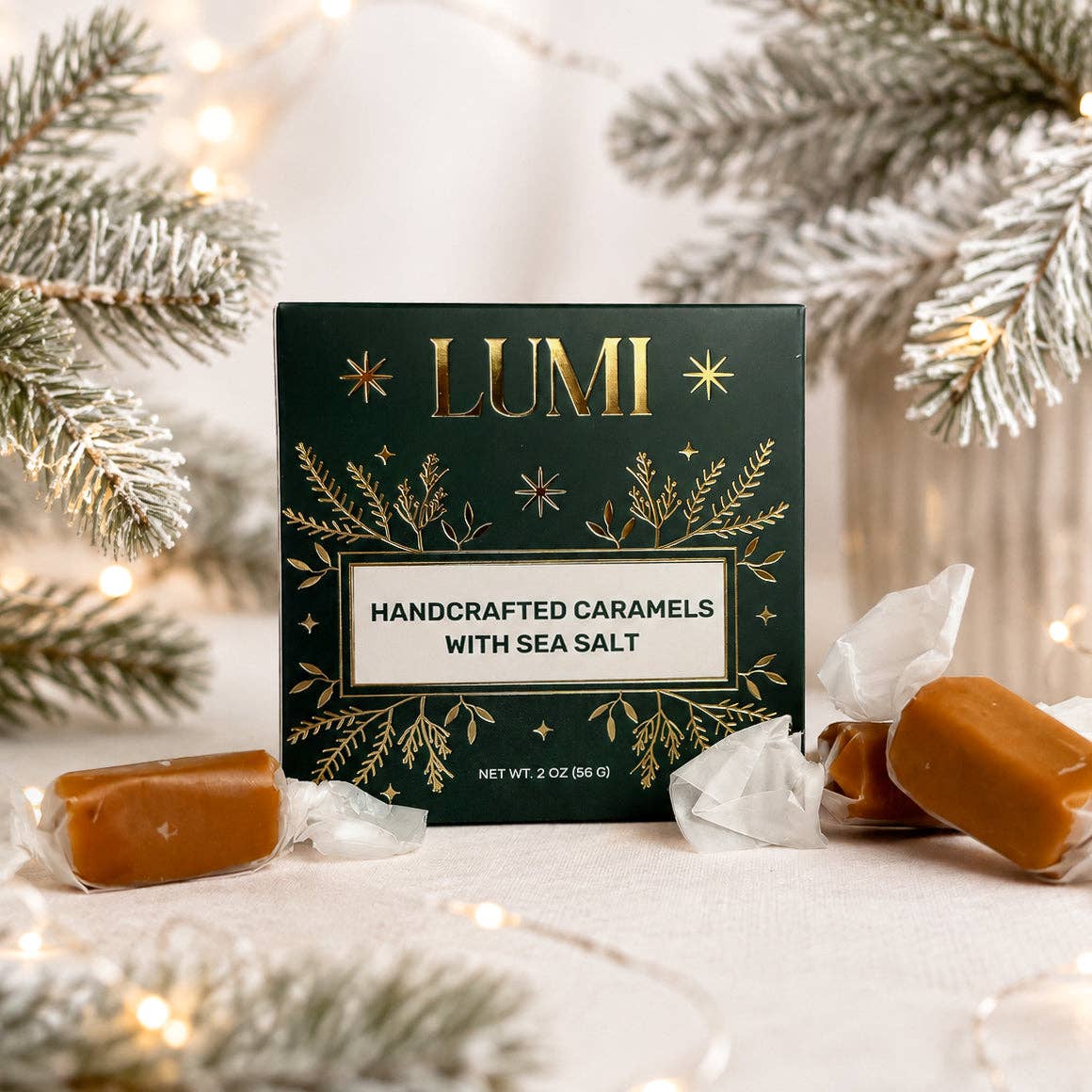 A box of LUMI Handcrafted Caramels with Sea Salt, styled with two unwrapped caramel candies on either side, set against a festive background of flocked pine branches and warm string lights.