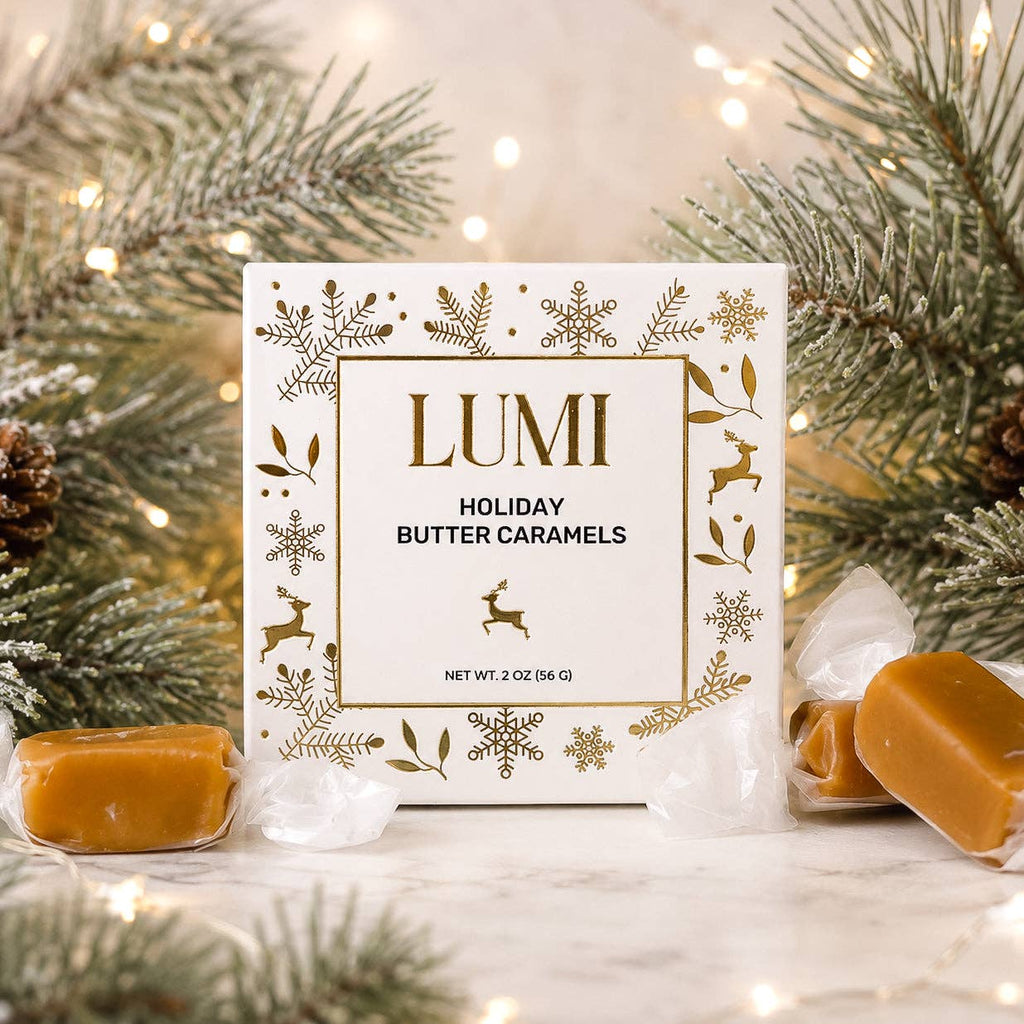 A boxed product labeled LUMI Holiday Butter Caramels sits on a marble surface surrounded by pine branches, warm string lights, and unwrapped caramel pieces. The box is white with gold decorative motifs and text, and the caramels are amber-brown.