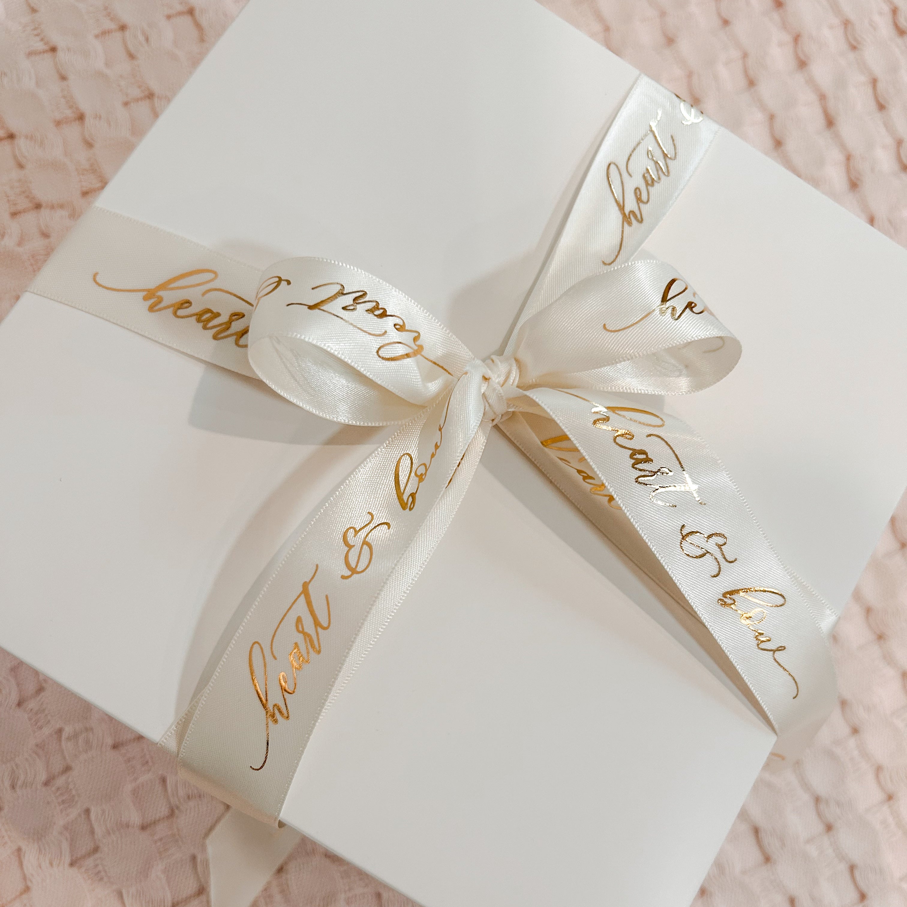 White gift box with gold text and a bow on a pink textured background