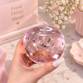 Translucent pink Chiboki Studio apple squishy filled with shimmering star confetti, held by a hand with white nails against a soft pink background with delicate flowers and gold accents.
