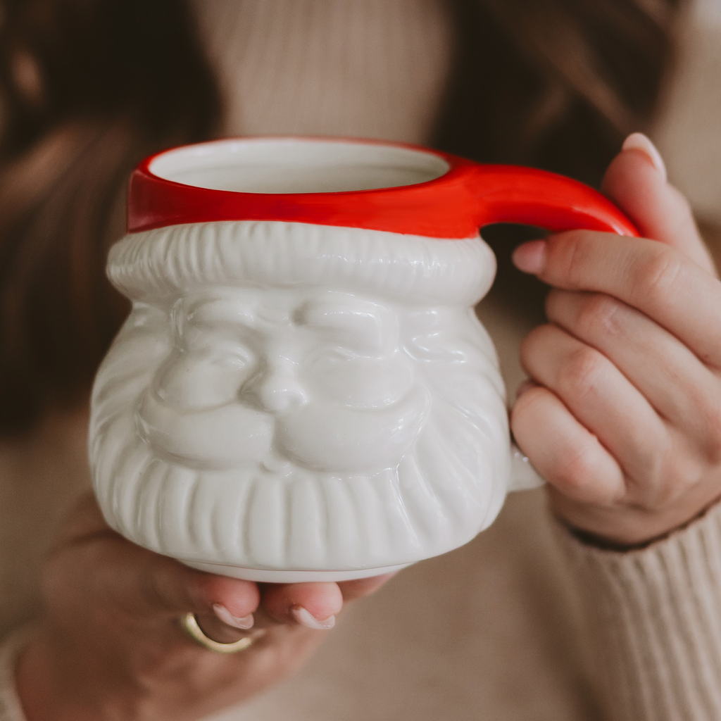 Cream ceramic Santa face mug with a bright red hat and curved red handle, held in two hands.

