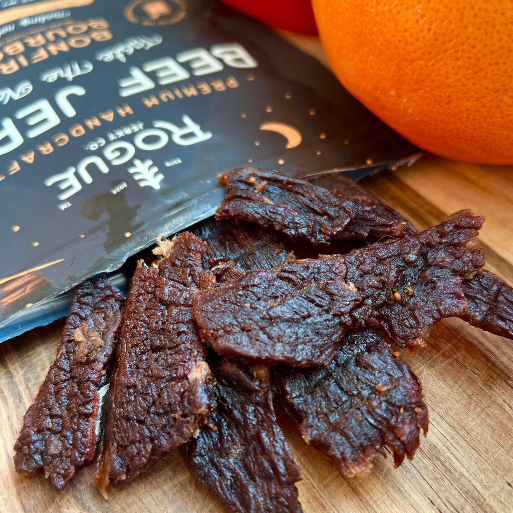Dried beef jerky on a wooden board with a Rogue Beef Jerky package and an orange in the background.