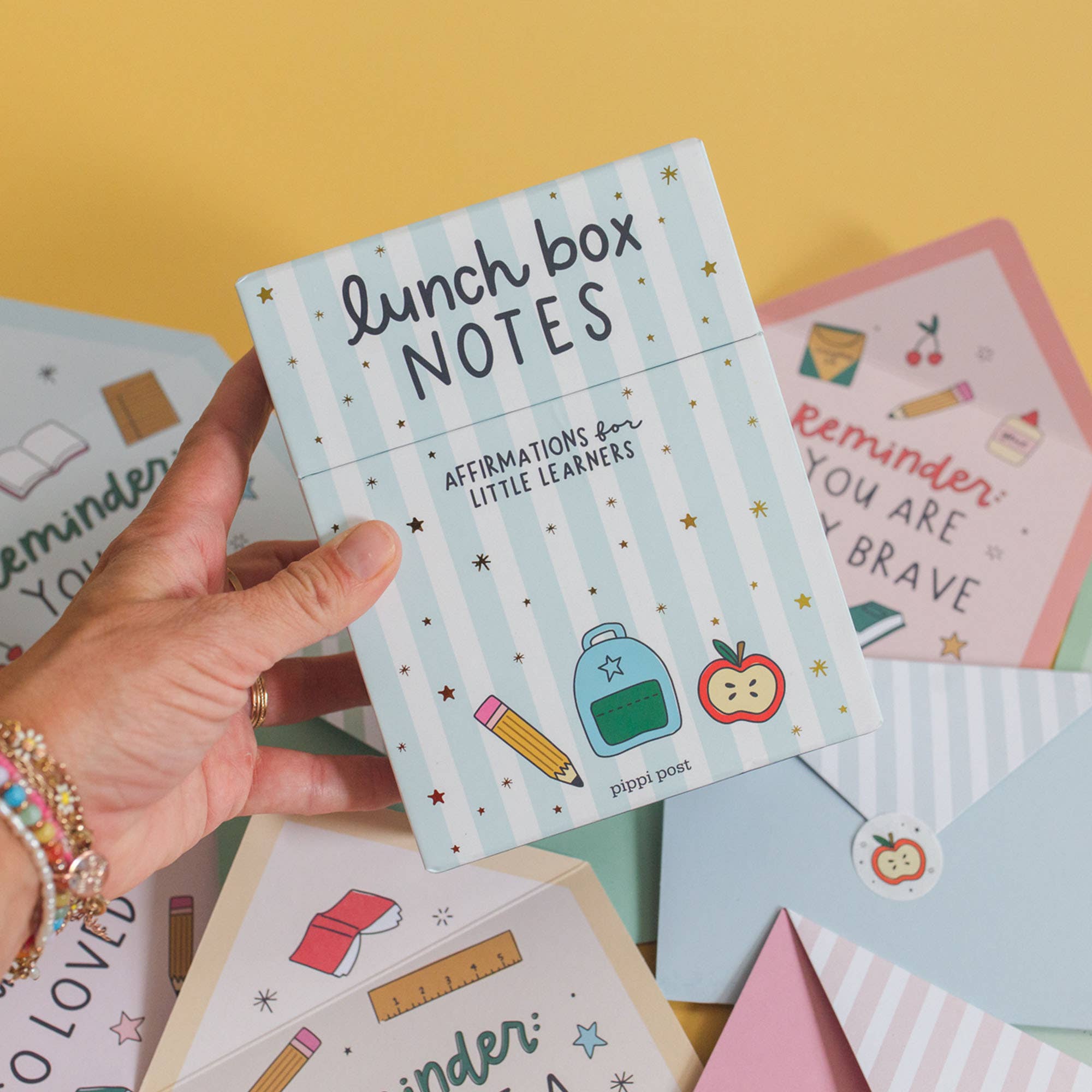 Hand holding a pack of 'lunch box notes' with colorful designs on a yellow background
