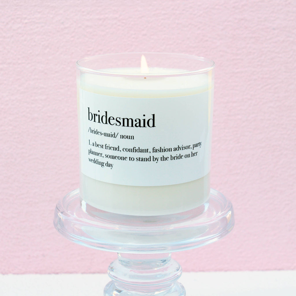 Bridesmaid Definition Soy Candle in a white glass jar with a sentimental definition-inspired label, displayed on a clear pedestal against a soft pink background.
