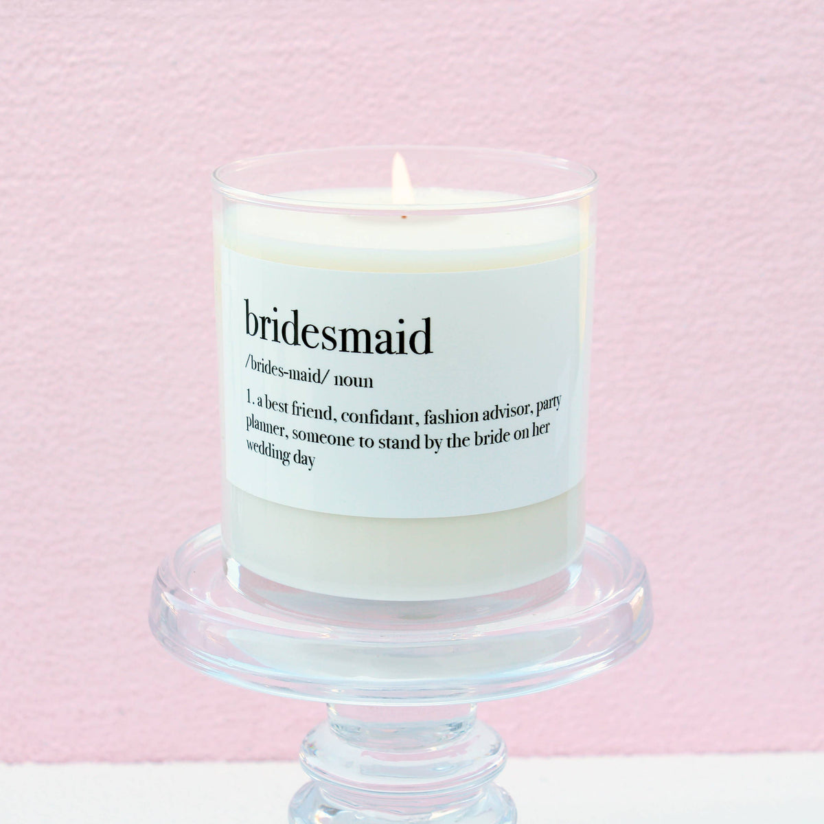 Bridesmaid Definition Soy Candle in a white glass jar with a sentimental definition-inspired label, displayed on a clear pedestal against a soft pink background.
