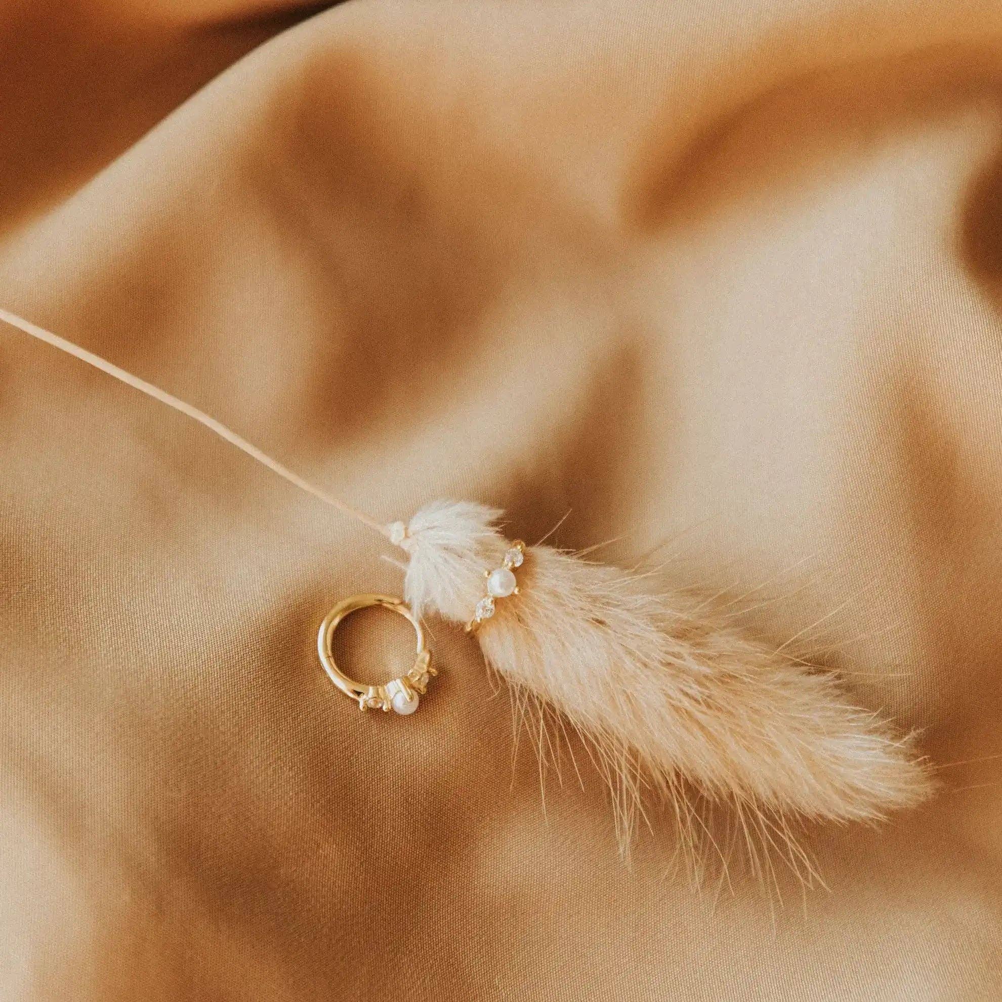 Gold ring with pearl on a string with dried pampas grass against a brown fabric background