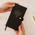 Hands holding a black faux leather Signature Edition pocket notebook with gold foil lettering and a snap button closure, perfect for work notes, journaling, travel, and everyday organization.
