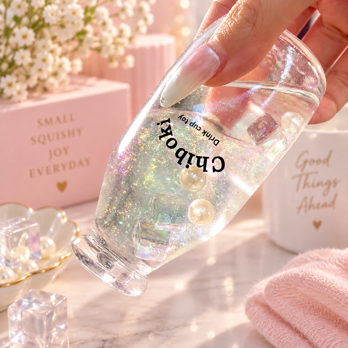 Chiboki Studio Crystal Clear Sand Bottle Squishy with iridescent glitter, pearl-like beads and clear cube details, held against a dreamy pastel pink background with delicate flowers and gold accents.