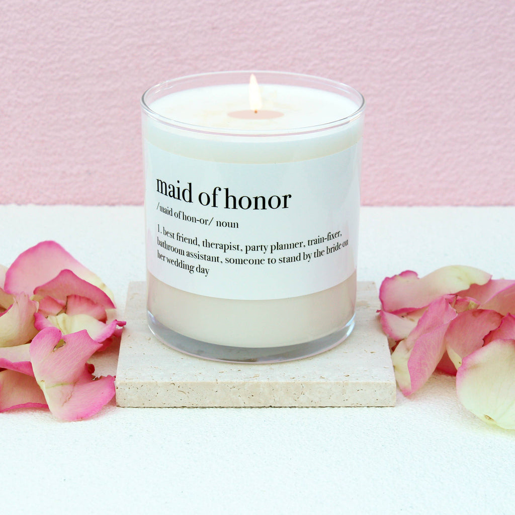A lit white candle in a clear glass jar sits on a small square stone coaster, surrounded by pink and white rose petals against a soft pink background. The label reads 'maid of honor' with a dictionary-style definition.