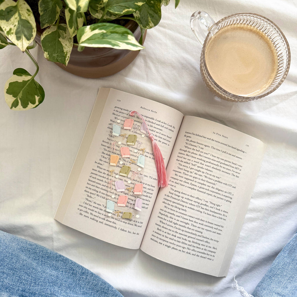 Open book with a pink bookmark, a cup of coffee, and a plant on a white surface.