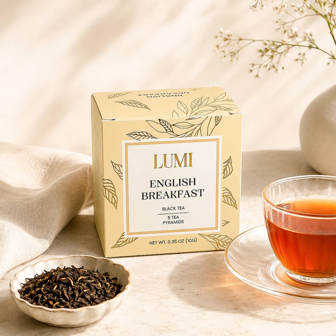 A box of Lumi English Breakfast black tea sits on a light beige surface beside a small dish of loose tea leaves and a clear glass cup of brewed tea, with soft natural shadows and a neutral decorative background.
