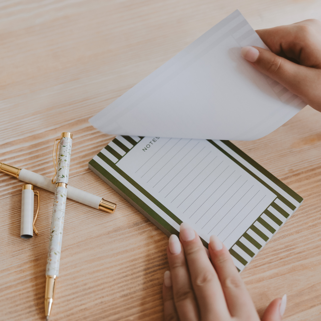 A sheet being lifted from a green striped lined notepad on a wooden desk beside white and gold pens.

