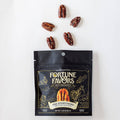 A black stand-up pouch of Fortune Favors Multicolor gourmet candy is shown on a white background with five pecans arranged above it. The pouch features gold decorative graphics and text, and the candy inside is partially visible through a clear window near the bottom.