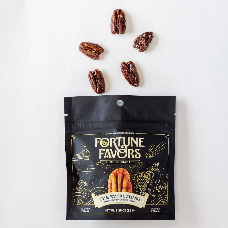 A black stand-up pouch of Fortune Favors Multicolor gourmet candy is shown on a white background with five pecans arranged above it. The pouch features gold decorative graphics and text, and the candy inside is partially visible through a clear window near the bottom.