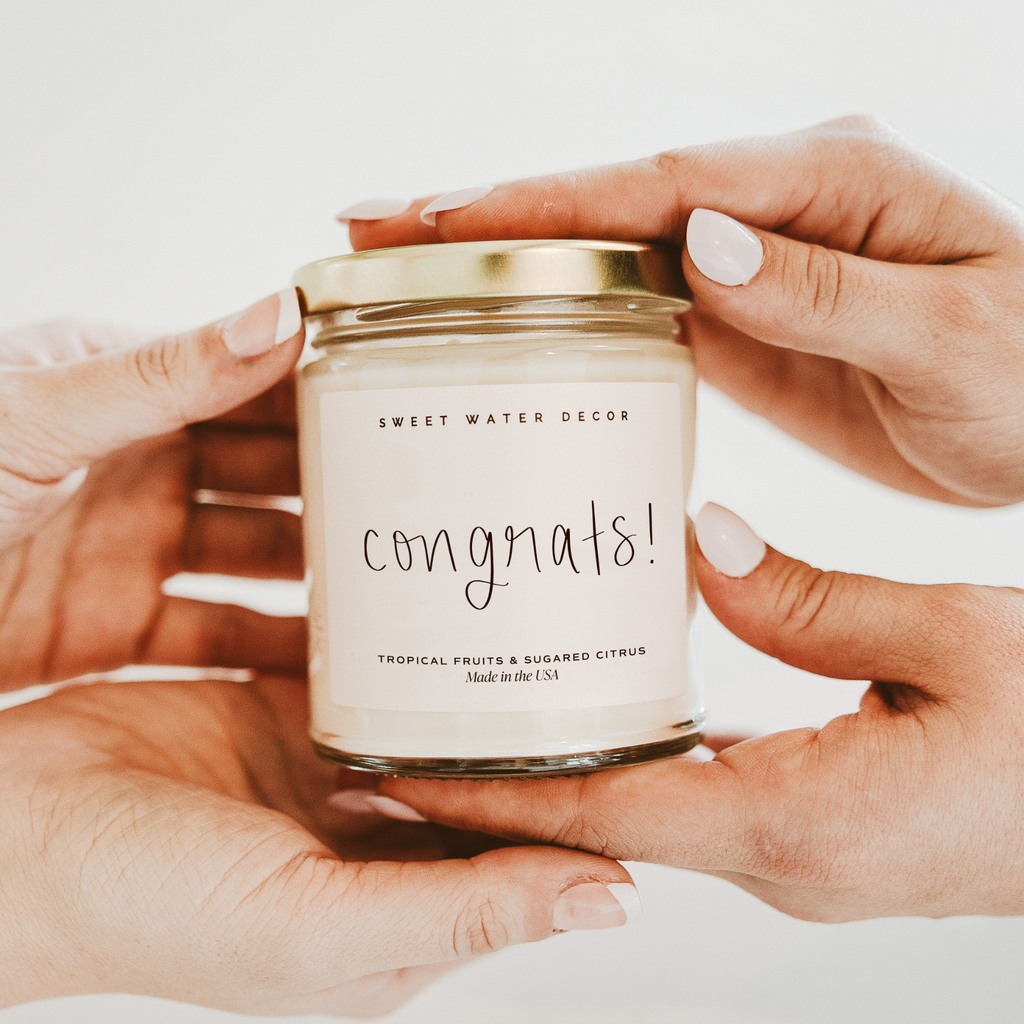 Candle jar labeled 'congrats!' held by hands against a neutral background