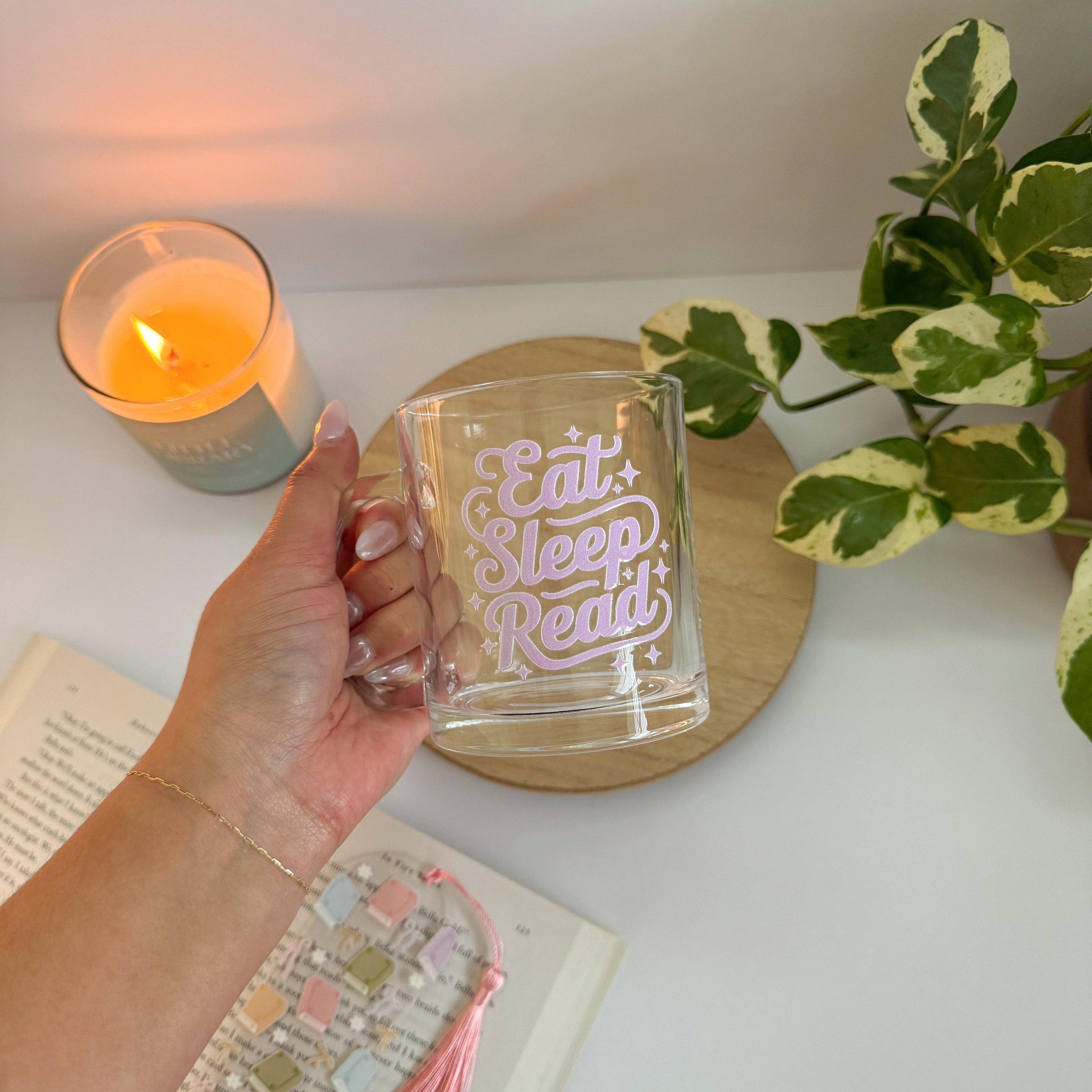 Hand holding a clear mug with 'Eat Sleep Read' text, candle, and plant in the background