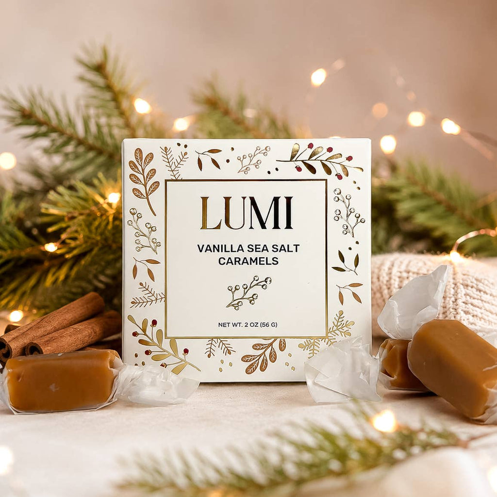 A boxed caramel candy product labeled LUMI Vanilla Sea Salt Caramels stands on a tabletop with wrapped caramels and cinnamon sticks around it, set against pine branches and warm string lights.