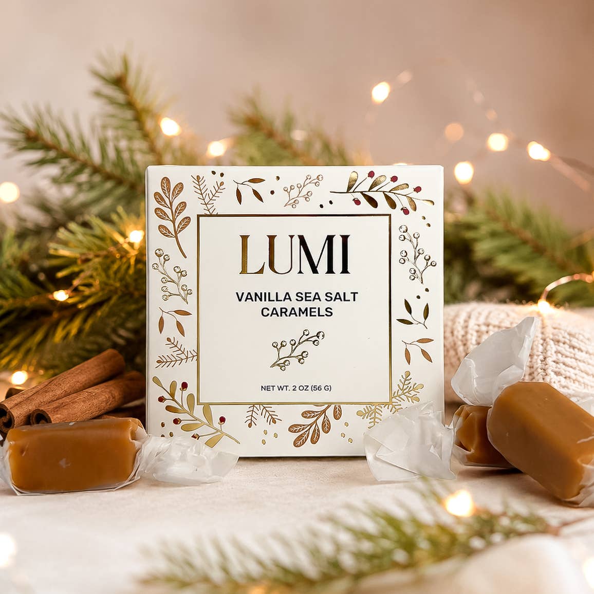 A boxed caramel candy product labeled LUMI Vanilla Sea Salt Caramels stands on a tabletop with wrapped caramels and cinnamon sticks around it, set against pine branches and warm string lights.