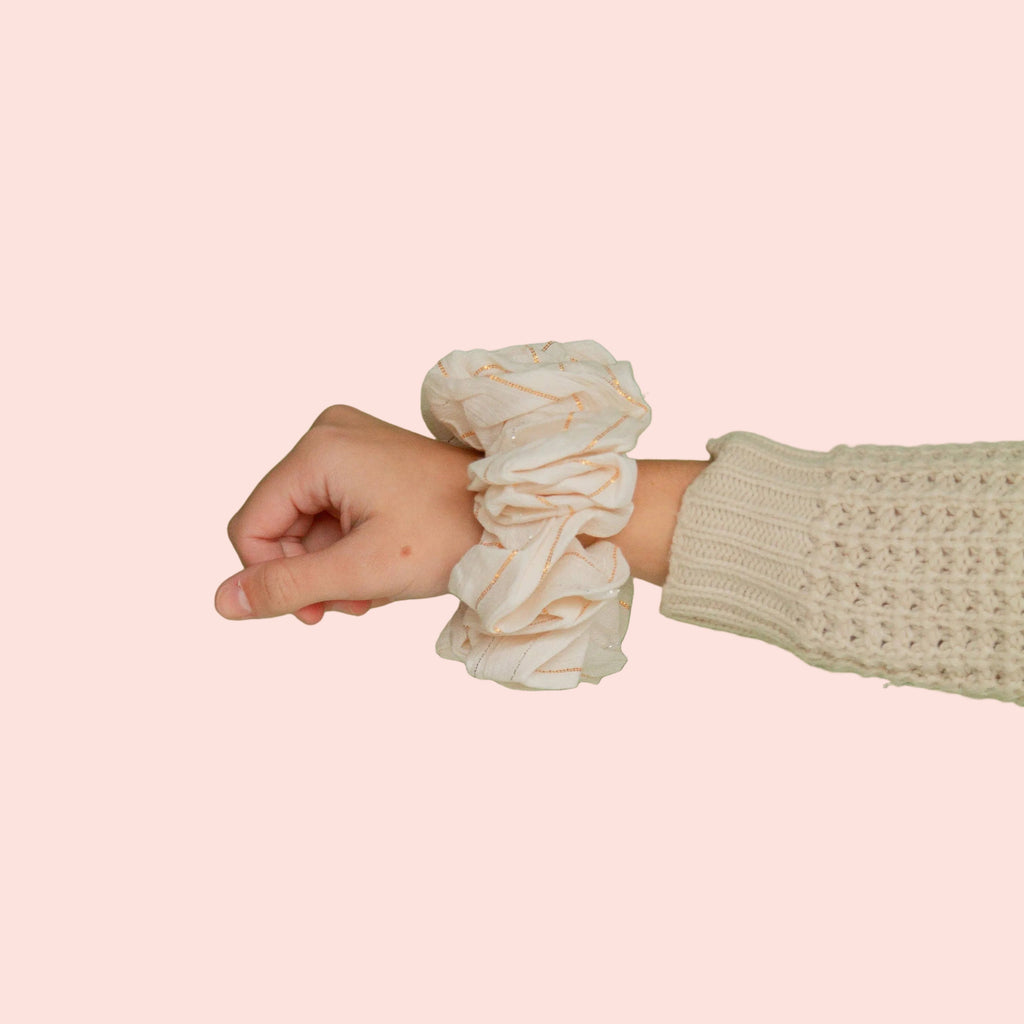 A cream-colored fabric scrunchie worn on a person's wrist, paired with a cream-colored knit sweater sleeve, set against a solid sage green background.