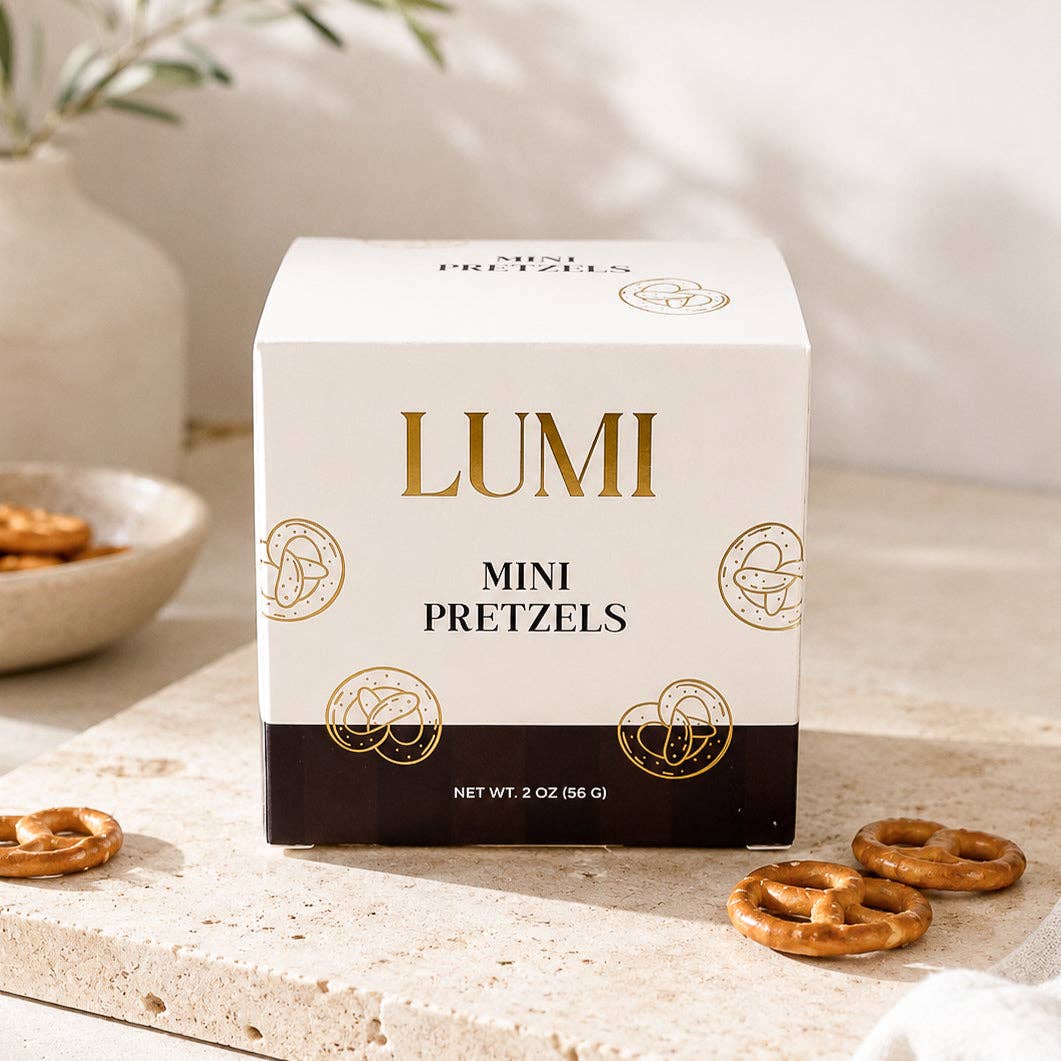 LUMI Mini Pretzels 2 oz box on a light stone surface with scattered pretzels, a snack bowl, and a vase in the background.
