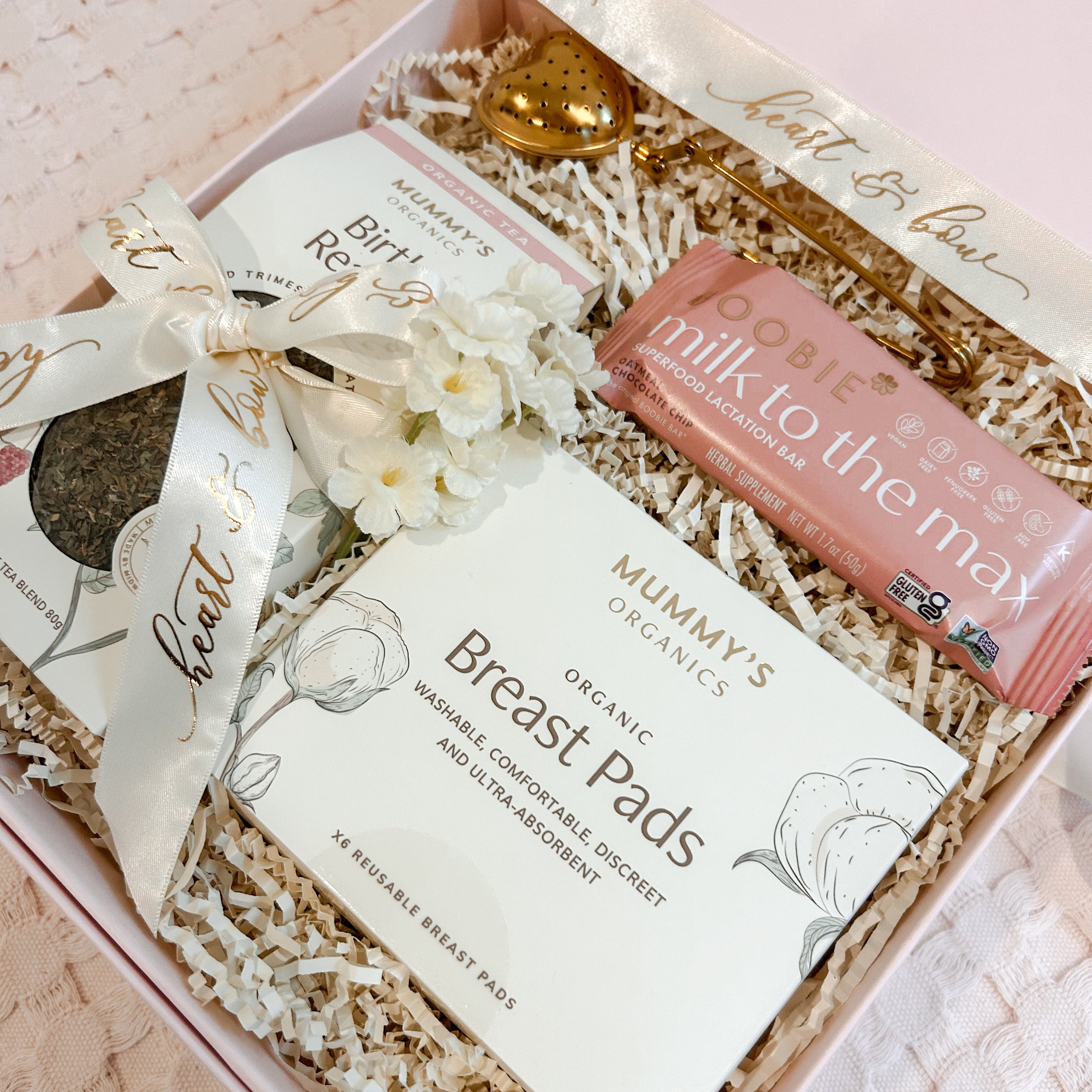 A curated gift box set containing Mummy's Organics Breast Pads, Boobie 'milk to, the max' bar, and a Birth Relief cream, arranged in a pink box with shredded paper filler and a gold teapot.