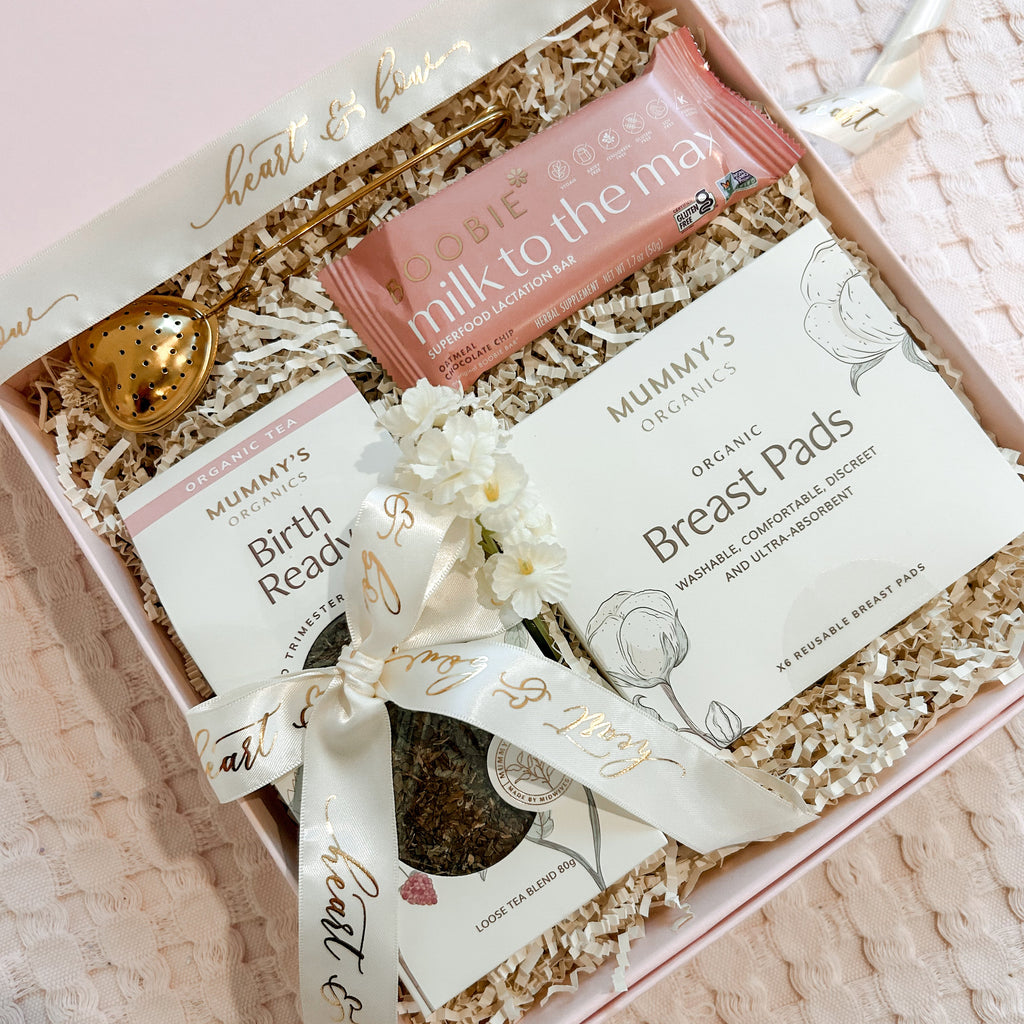 A pink gift box filled with breastfeeding support items, including Mummy's Organics Breast Pads, a 'milk to the may' breast pump battery, a gold heart-shaped egg, and a small white flower, tied with a white ribbon printed with 'heart to heart'.