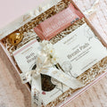 A pink gift box filled with breastfeeding support items, including Mummy's Organics Breast Pads, a 'milk to the may' breast pump battery, a gold heart-shaped egg, and a small white flower, tied with a white ribbon printed with 'heart to heart'.