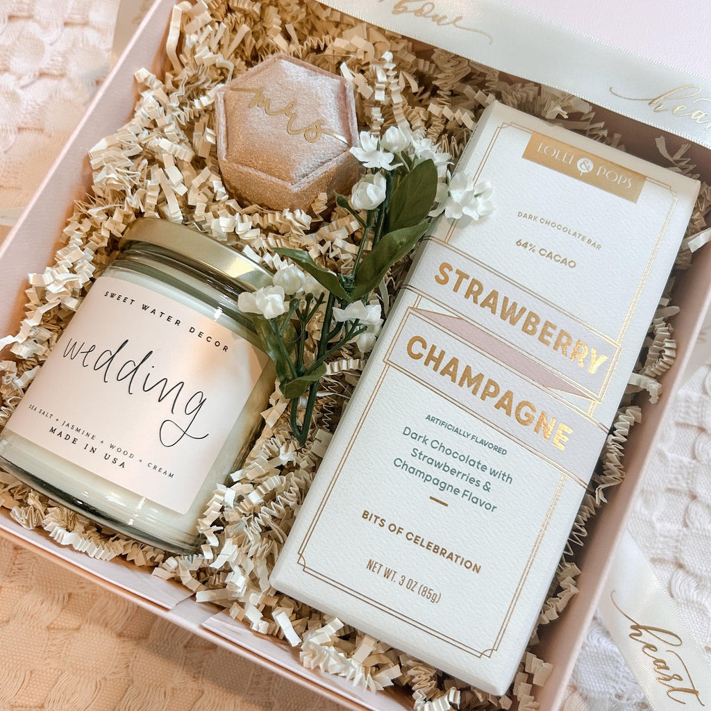 Gift box with 'wedding' candle, chocolate bar, and flowers on a light background