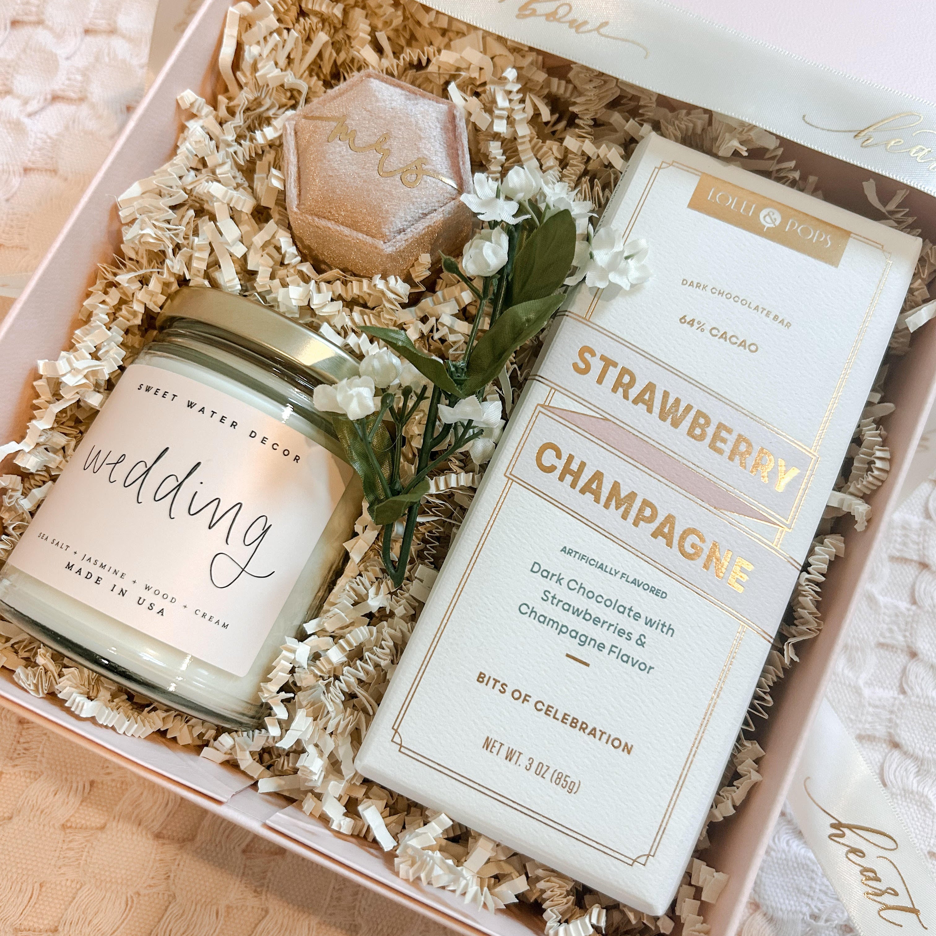 Gift box with 'wedding' candle, chocolate bar, and flowers on a light background