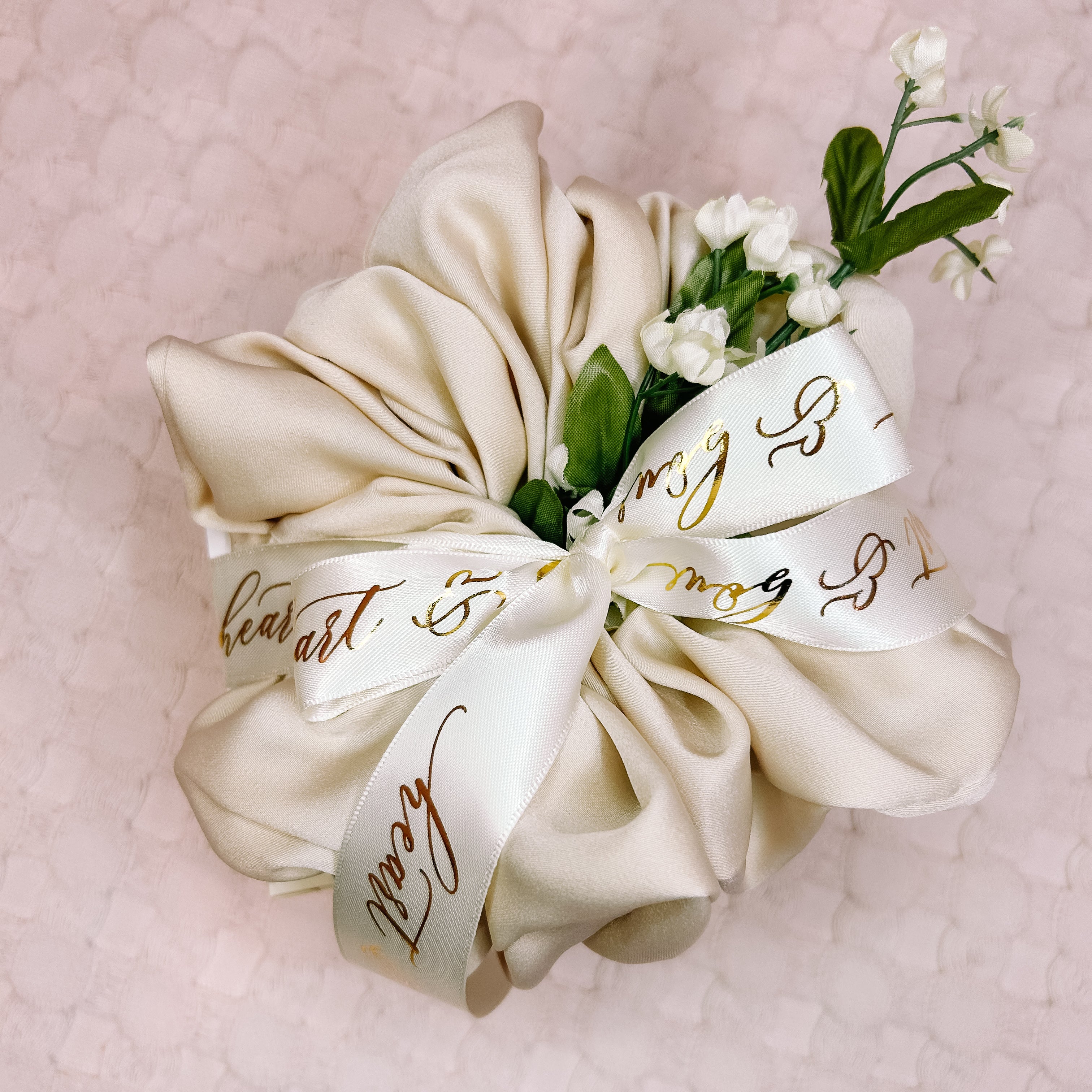 Cream-colored scrunchie with floral accents and gold text on a pink textured background