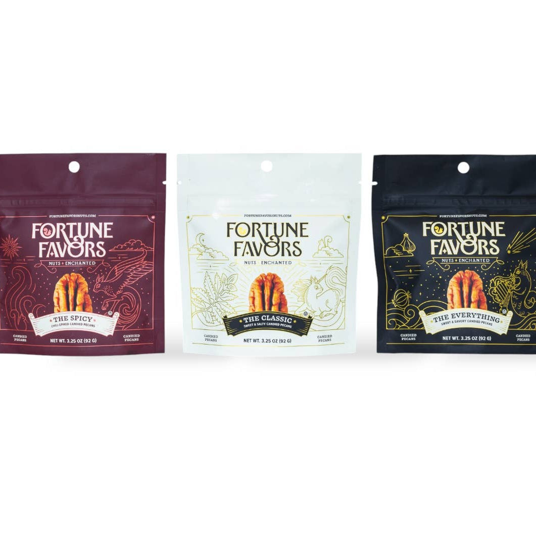 Three stand-up pouches of Fortune Favors candy arranged side by side on a light gray background. The left pouch is dark burgundy with gold and white text, the middle pouch is white with pale gold text, and the right pouch is black with gold text.
