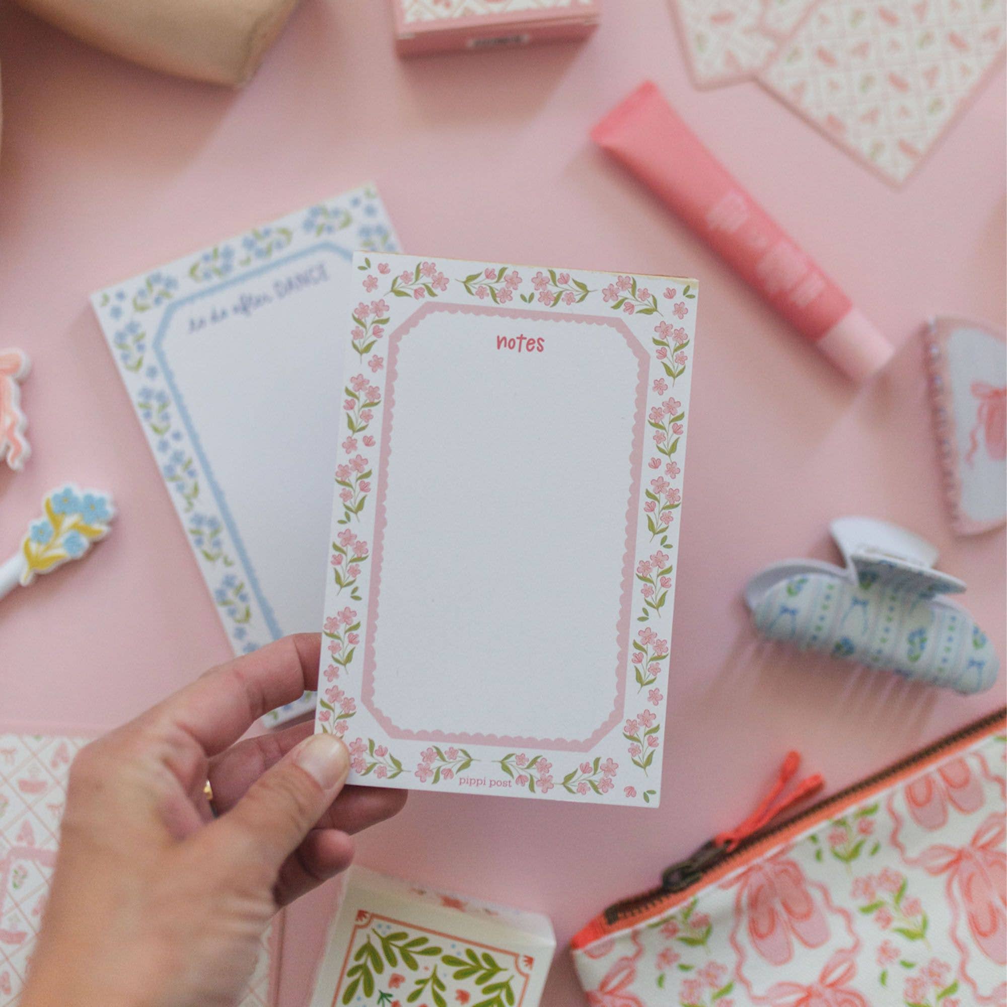 A hand holds a white floral notepad with a pink border and the word 'notes' at the top, surrounded by matching pastel stationery items on a pink surface.
