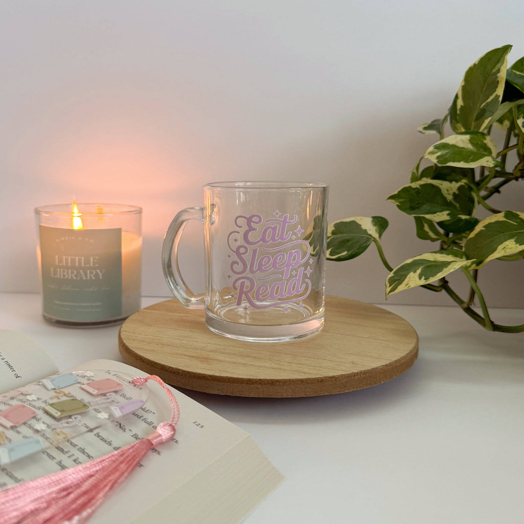 Glass mug with 'Eat Sleep Read' text on a wooden coaster next to a candle and book.