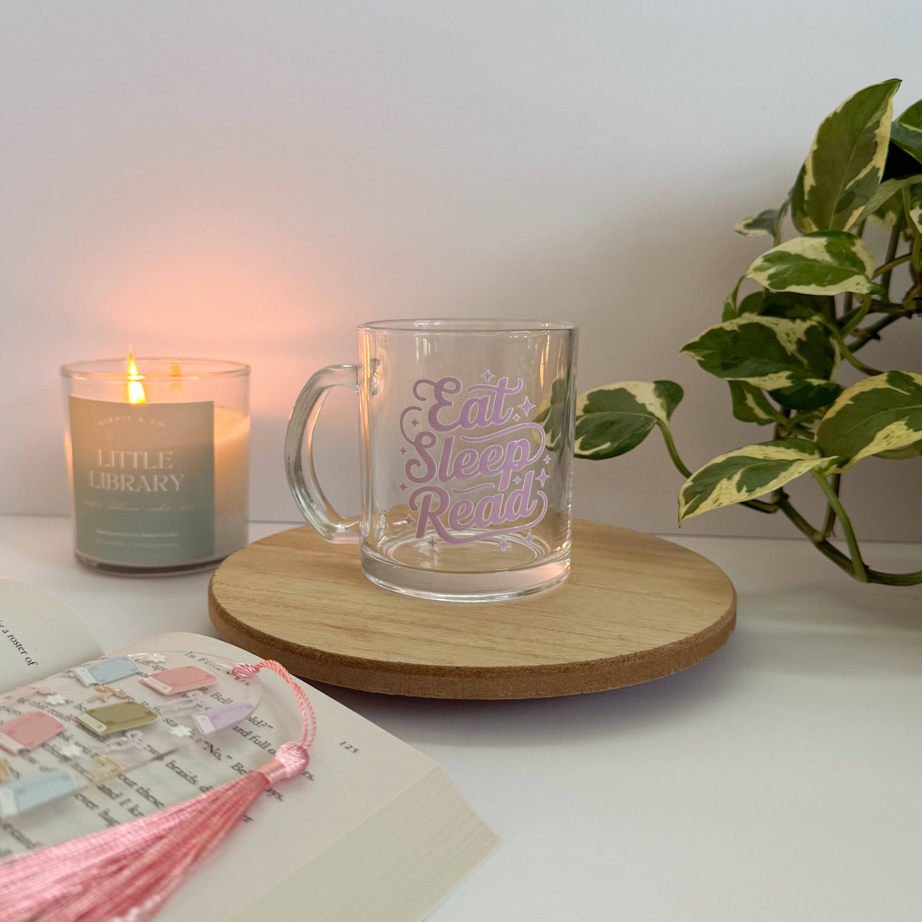 Glass mug with 'Eat Sleep Read' text on a wooden coaster next to a candle and book.