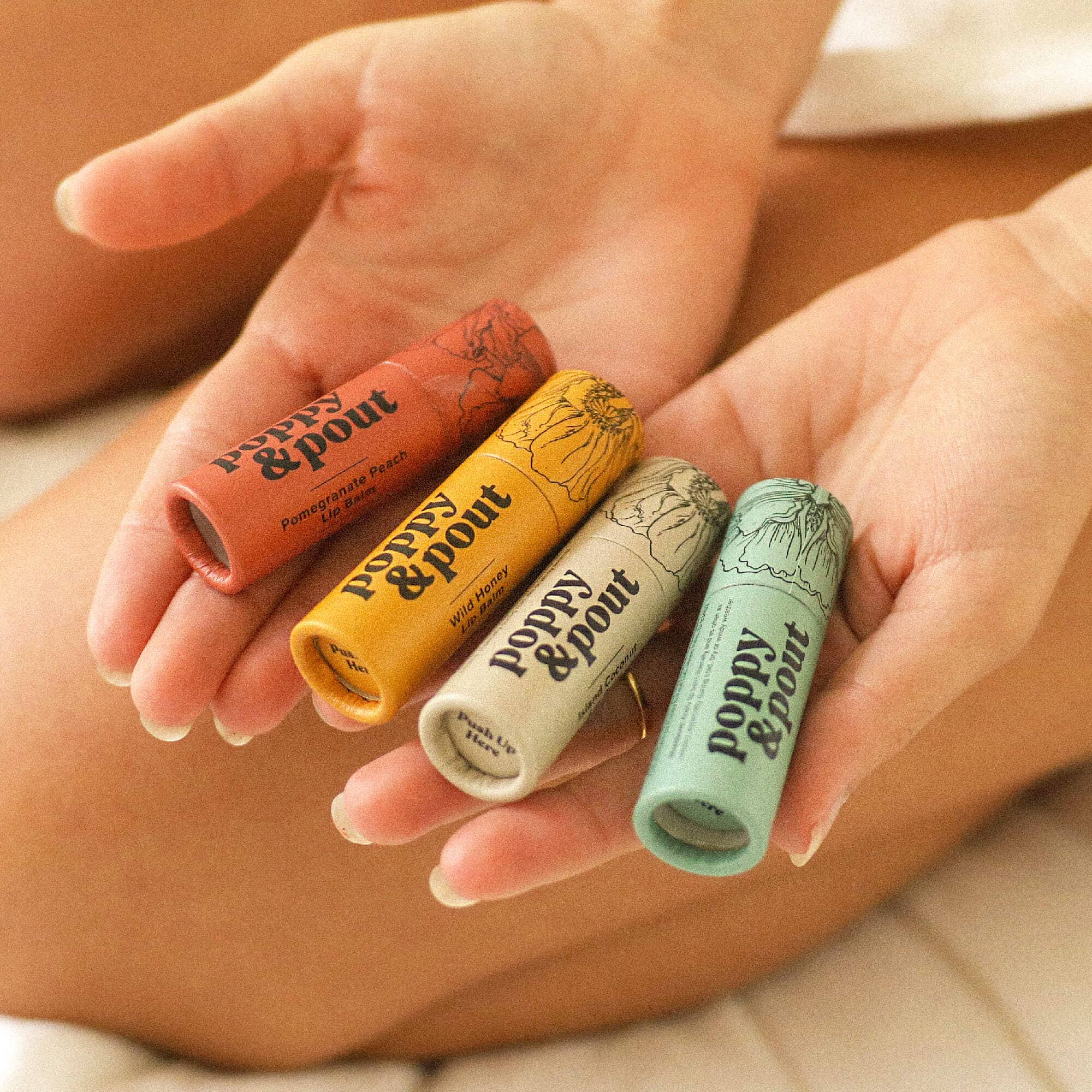 Four lip balm tubes from Poppy & Pout held in a hand against a neutral background