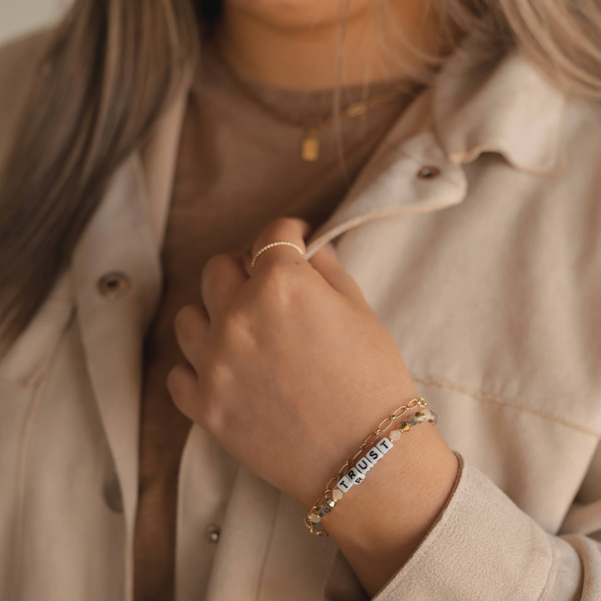 Person wearing a beige jacket with a close-up of their hand and jewelry.