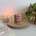 Glass mug with 'Forget flowers, buy me books' text on a wooden coaster with a candle and plant in the background.