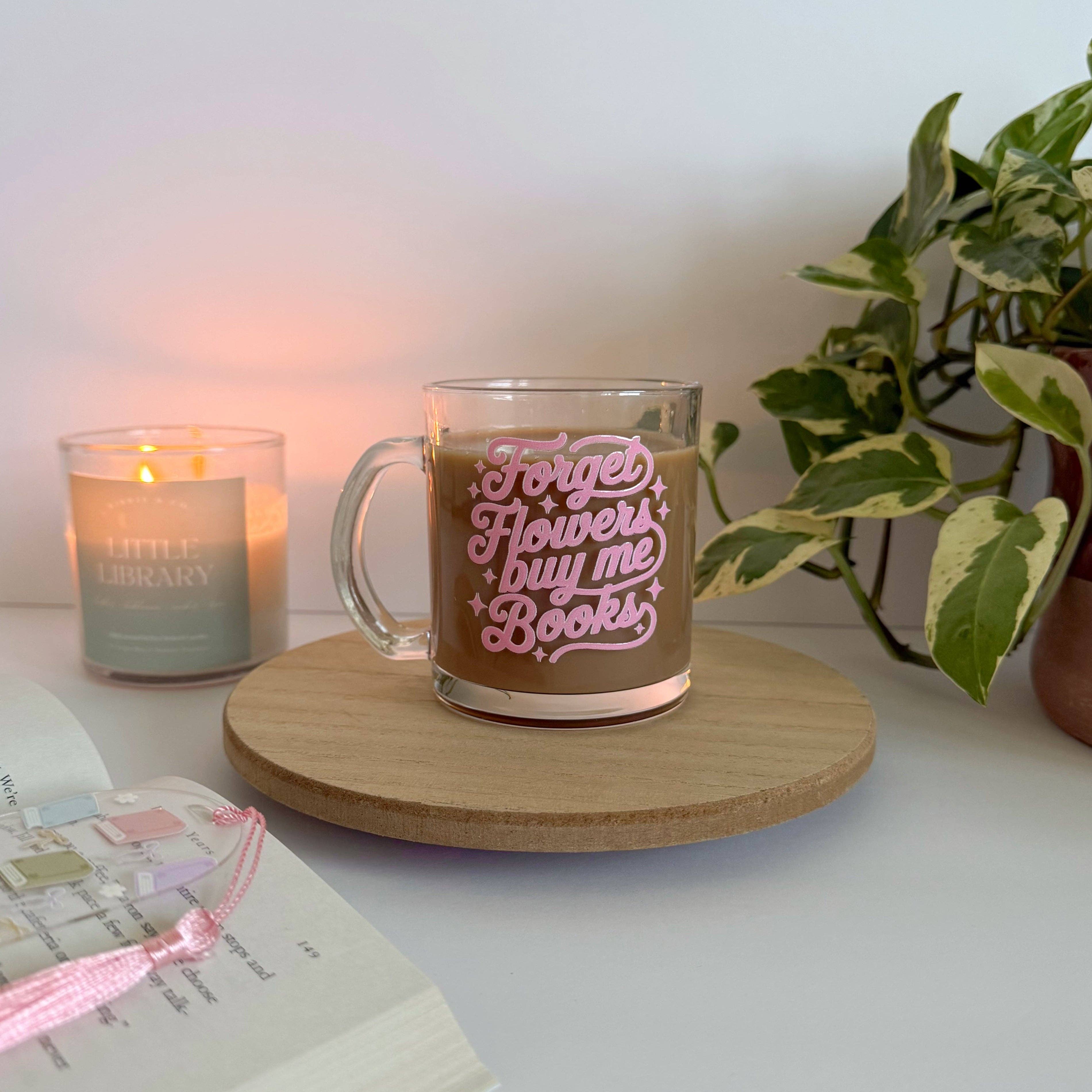 Glass mug with 'Forget flowers, buy me books' text on a wooden coaster with a candle and plant in the background.