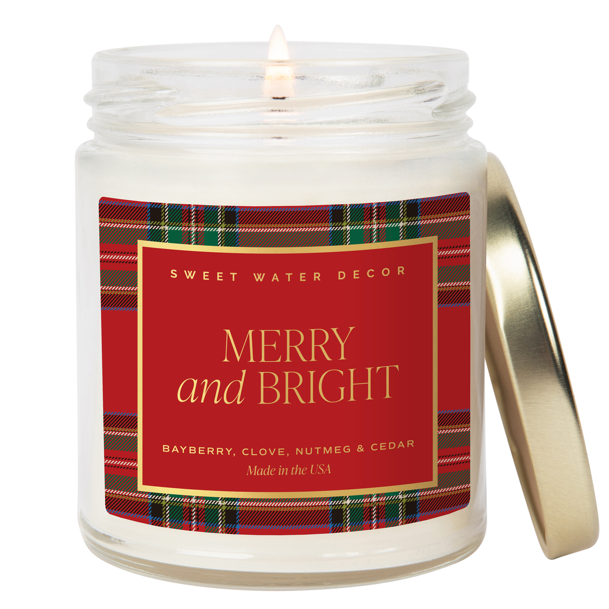 Lit Merry & Bright Christmas candle with a red and green plaid label and a gold-tone lid resting beside the clear glass jar.