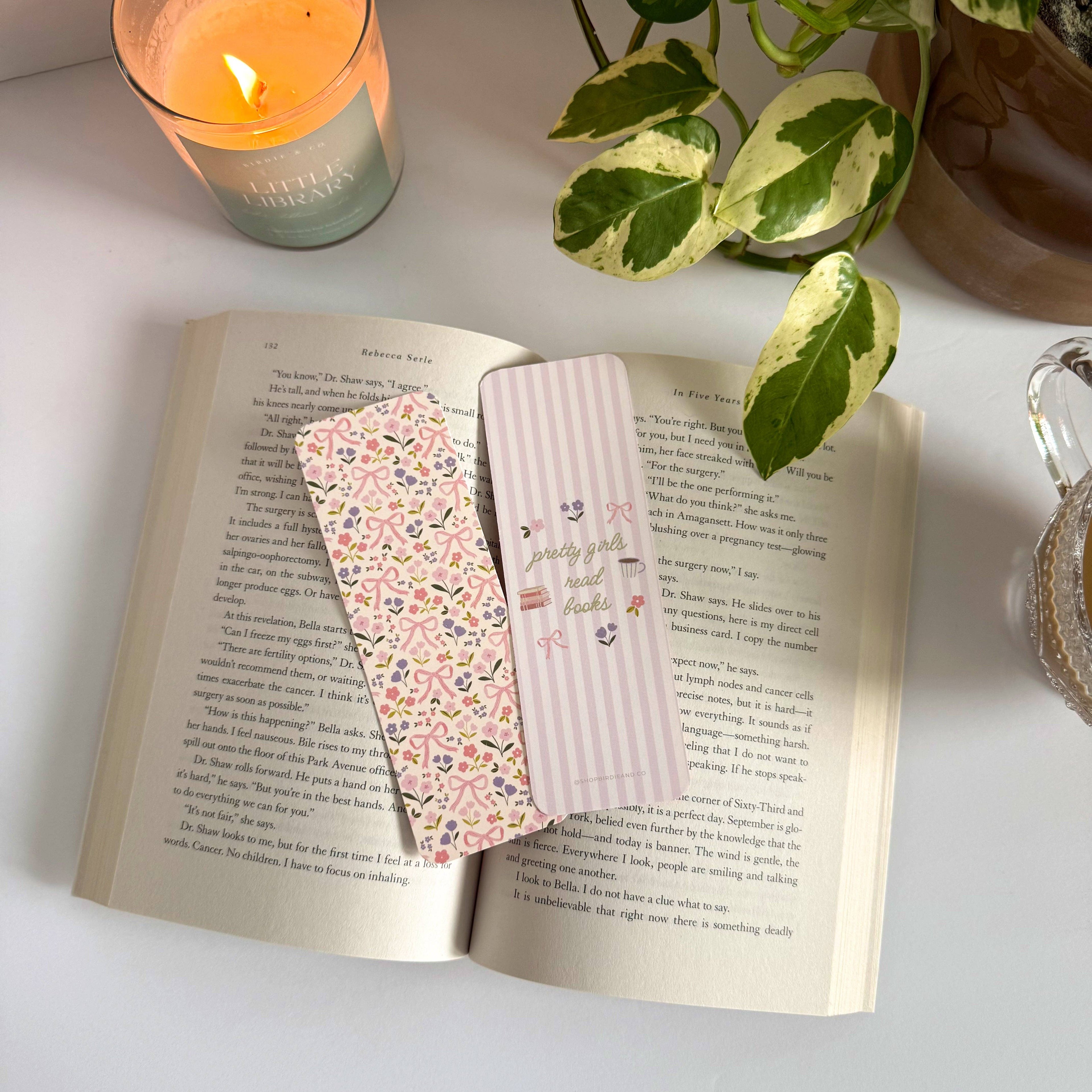 Open book with floral and striped bookmarks on a light surface with a candle and plant in the background.
