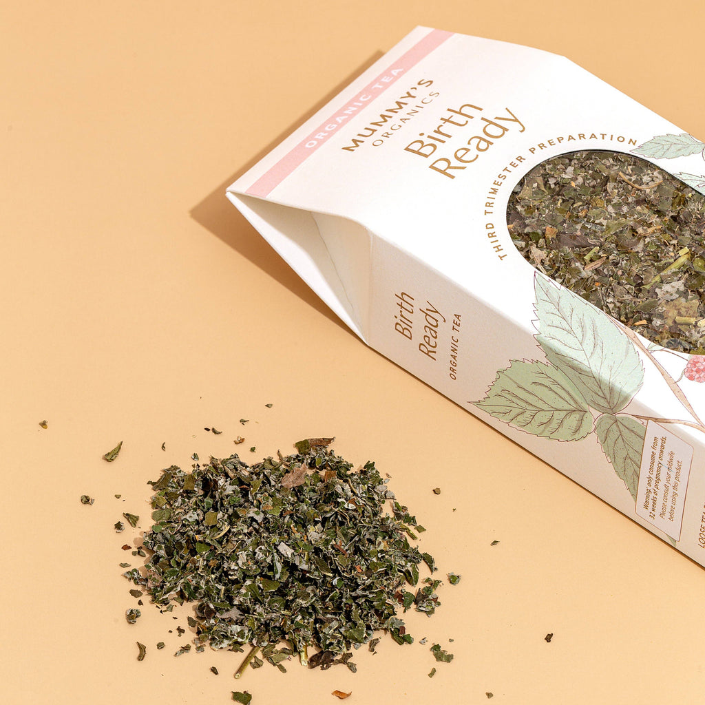 A package of Mummy's Organics Birth Ready Organic Tea lies on a warm beige background with a small pile of loose green tea leaves in front. The white and pink box features a clear window showing the dried leaf blend inside.