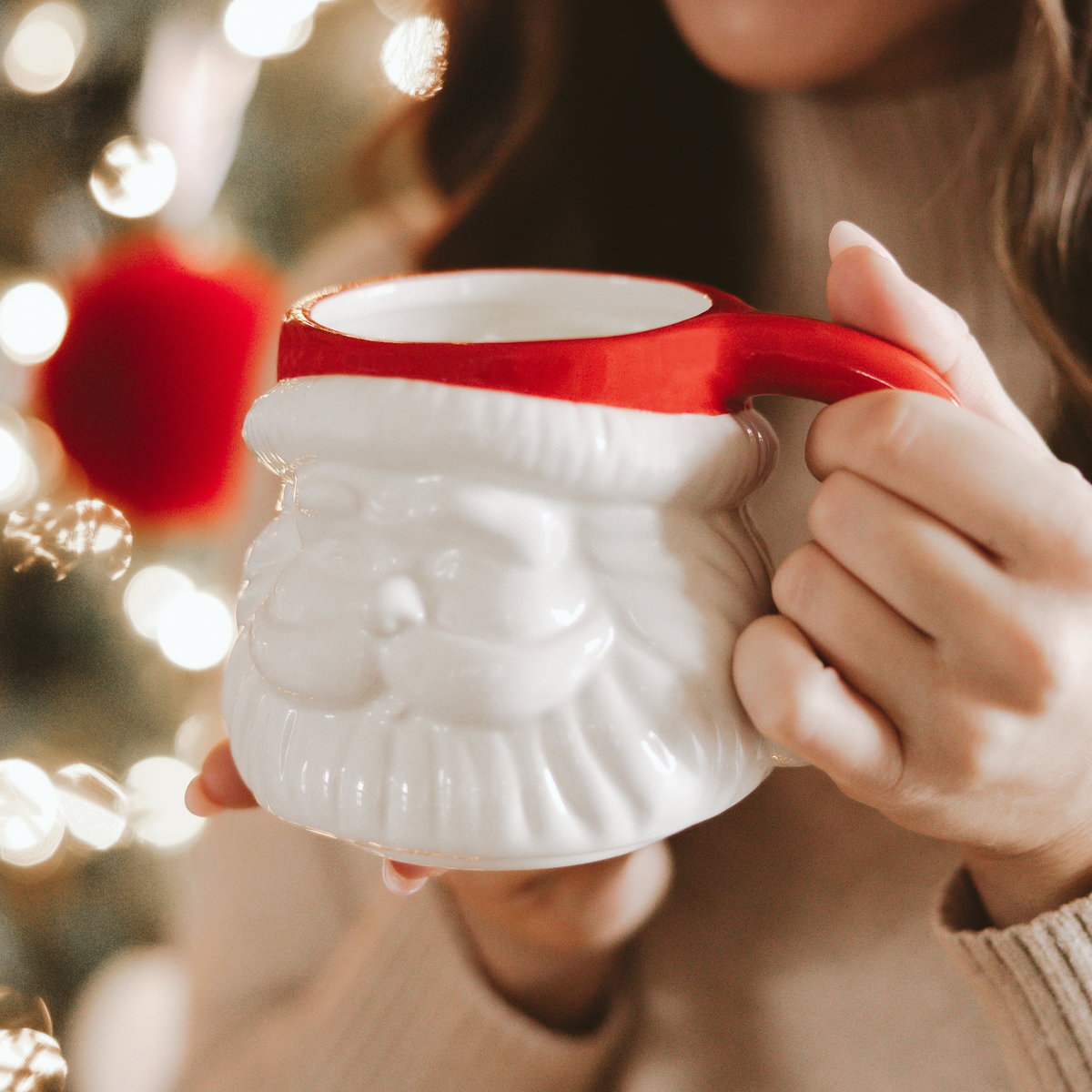 Person holding a cream Santa face mug with a red hat and handle beside a Christmas tree with glowing lights.
